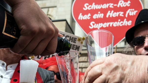 Im Hintergrund ein Schild "Schwarz Rot hat ein Herz für Superreiche"- Demonstranten halten am 30.06.2016 vor dem Bundesrat in Berlin Einweggläser in den Händen und lassen sich Sekt einschütten. Die Organisation Attac hatte zu einer Protestaktion gegen das geplante Erbschaftssteuer-Gesetz aufgerufen.  | Bild: picture-alliance/dpa