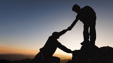 Eine Person reicht einer anderen Person die Hand, um ihr zu helfen, einen Fels hinaufzuklettern. | Bild: Colourbox.com/Tinnakorn Jorruang