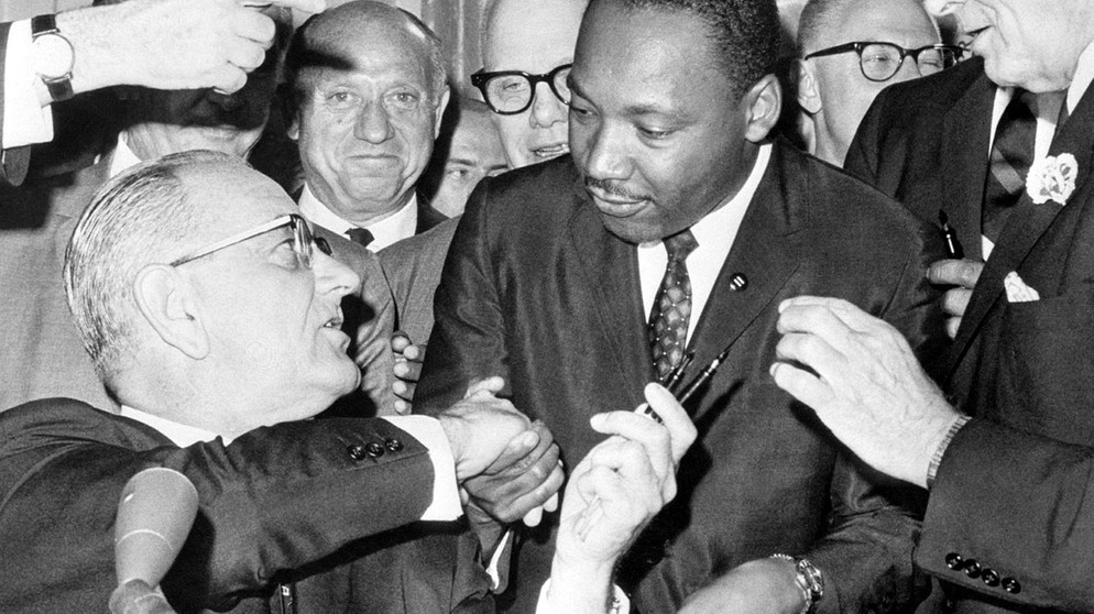 Nach der Unterzeichnung des Bürgerrechts-Gesetzes am 2.7.1964 in Washington DC. reicht US-Präsident Lyndon Baines Johnson (l) den Federhalter an den Bürgerrechtler Martin Luther King weiter und gibt ihm die Hand. | Bild: picture-alliance/dpa
