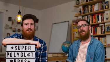 Die superpolyglotten Zwillinge Matthew und Michael Youlden mit einem Schild auf dem "Super Polygl@t Bros" steht. Im HIntergrund sind regale zu sehen | Bild: picture-alliance/dpa