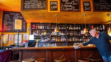 Matt wischt den hölzerenen Tresen seiner Bar ab. im Hintergrund stehen viele Flaschen in einem Holzregal.  | Bild: picture alliance / SZ Photo 