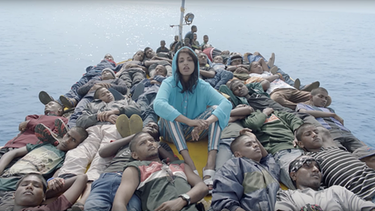 Frau in blauem Kaputzenpulle sitzt in einer Mitte eines vollen Flüchtlingsboot, die jungen Männer um sie herum liegen. | Bild: Screenshot: M.I.A. - "Borders"