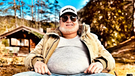 Stefan Raab posiert mit Bierbauch (Fatsuit oder KI-generiert?) und Kappe mit der Aufschrift "NWSDWH" vor einer hügeligen, bewaldeten Landschaft. Er trägt eine Pilotensonnenbrille und einen grauen drei Tage Bart.  | Bild: therealstefanraab / Instagram