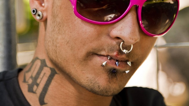 Ein junger Mann mit Piercings in Ohr, Nase, Ober- und Unterlippe und einer Tätowierung am rechten Hals. | Bild: picture-alliance/dpa