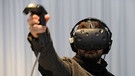 Mann trägt Datenbrille, in der Hand einen Joystick, den er wie eine Waffe mit ausgestrecktem Arm einsetzt | Bild: picture-alliance/dpa