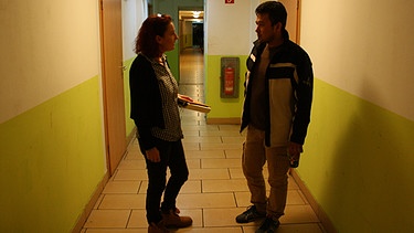 Ein junger Asylbewerber spricht mit einer rothaarigen Frau auf einem Gang von dem viele Türen weggehen, der Boden ist gekachelt | Bild: BR/Wolfgang Kerler