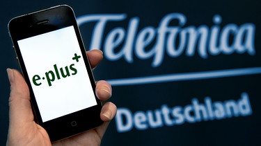 Eine Hand hält ein iPhone mit dem Logo von E-Plus auf dem Bildschirm vor dem Hintergrund des Logos von Telefónica Deutschland | Bild: picture-alliance/dpa