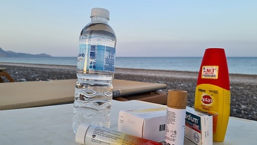 Ein Strand in der Abendstimmung. Im Vordergrund Medikamente, Insektenschutz und Mineralwasser - ein Symbolbild für die Reiseapotheke im Urlaub. | Bild: picture-alliance/dpa