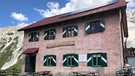 Berglhütte: Ein einfacher Bau, rosafarbene Fassade, grün-weiße Fensterläden. Wuchtige Holztische davor. | Bild: BR/Kilian Neuwert