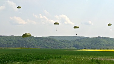 Mehrere Fallschirmspringer befinden sich in der Luft über einer grünen Landschaft | Bild: BR