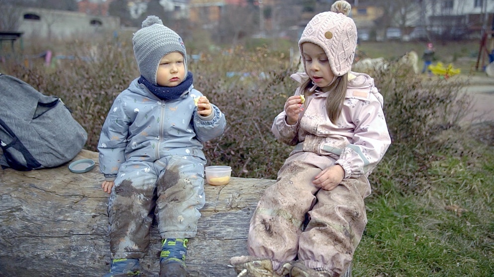 Zwei Kinder in Winterkleidung mit Mützen essen auf einem Baumstamm. | Bild: BR