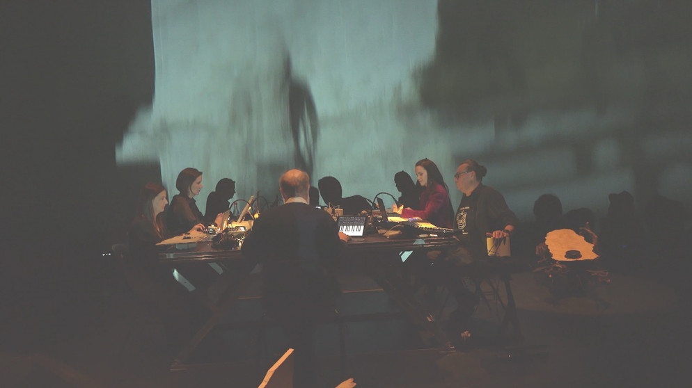 Personen mit Geräten an einem Tisch im Halbdunklen | Bild: BR