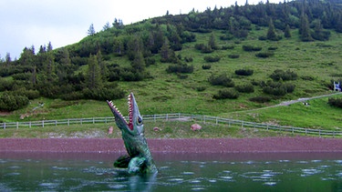 Ein Dinosauriermodell ragt aus einem Speichersee in Österreich | Bild: BR