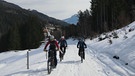 Drei Mountainbiker mit Skiern auf den Rucksäcken fahren durch ein verschneites Tal | Bild: BR