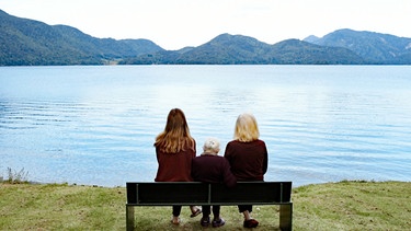 Drei Frauen aus unterschiedlichen Generationen sitzen auf einer Bank mit dem Rücken zum Betrachter und schauen auf den Walchensee | Bild: BR