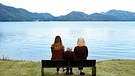 Drei Frauen aus unterschiedlichen Generationen sitzen auf einer Bank mit dem Rücken zum Betrachter und schauen auf den Walchensee | Bild: BR