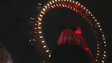 Frau mit Mickey Mouse Ohren und einer pinken Perücke vor einem Riesenrad | Bild: BR