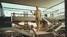 Eine riesige, nackte ältere Frau, in mitten von einer Bibliothek mit vielen irreführenden Treppen | Bild: BR