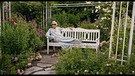 Ältere Person, welche auf einer weißen Parkbank, in Mitten von einem Garten posiert. | Bild: BR