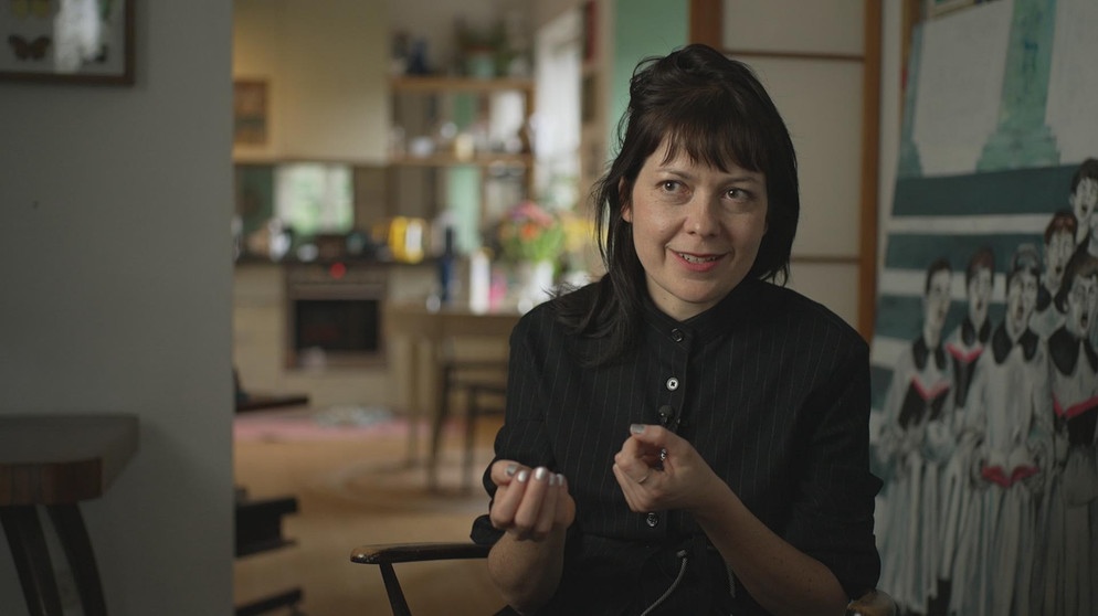 Eine Frau (Dana von Suffrin) mit schwarzem Haar, welche in mitten einer Wohnung sitzt und redet. | Bild: BR