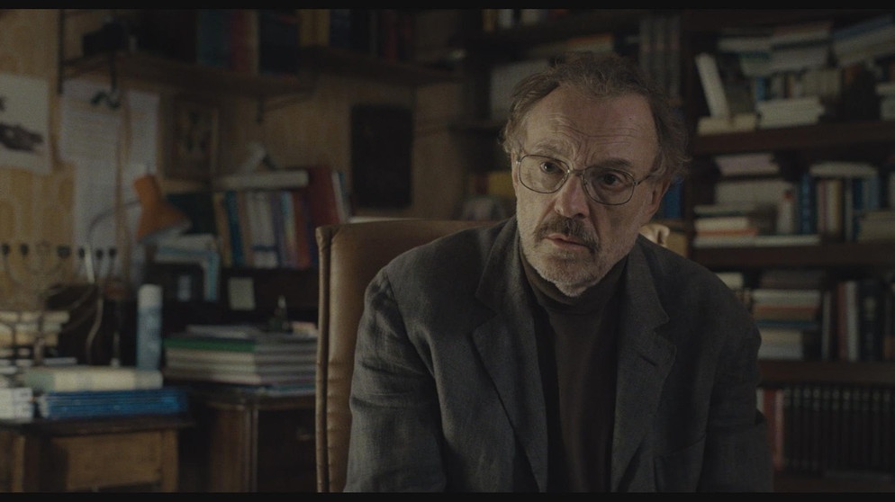 Ein Mann (Josef Hader) sitzt vor der Kamera, hinter ihm ein holziger Raum mit einem Regal voller Bücher. | Bild: BR