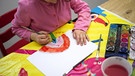 Ein Kind, an einem Tisch, welches mit Wasserfarben einen Regenbogen malt. | Bild: BR