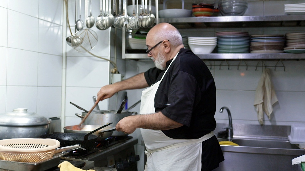 Ein Mann mit einem großen Bauch und einer Schürze kocht in einer Küche. | Bild: BR