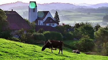 Eine Kirche und Häuser in hügeliger Landschaft | Bild: BR