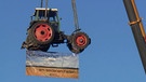 Ein Traktor wurde von einem Kran in die Höhe gezogen. An diesem hängt ein Transparent mit der Aufschrift "Landwirtschaft am seidenen Faden". | Bild: BR