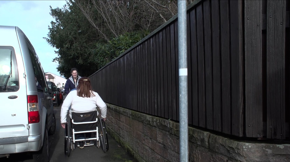 Eine Rollstuhlfahrerin fährt einer anderen Frau in einem engen Gehsteig entgegen. | Bild: BR