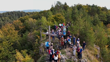 Menschen stehen auf einem bewaldeten Berg mit Gipfelkreuz | Bild: BR Fernsehen