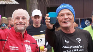 Zwei Sportler halten Getränke in die Kamera und lächeln dabei. | Bild: BR
