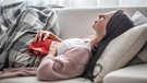 Frau liegt mit einer karierten Decke und einer roten Wärmflasche auf einem Sofa. Sie leidet unter Bauchschmerzen. | Bild: Colourbox