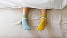 Jemand trägt im Bett zwei verschiedenfarbige Socken. | Bild: picture-alliance/dpa
