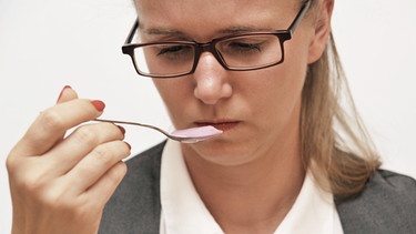 Eine junge Frau mit Brille hält einen Löffel vor den Mund und macht dabei einen skeptischen Gesichtsausdruck | Bild: picture alliance / Romain Fellens