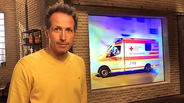 "Gut zu wissen"-Moderator Willi Weitzel mit Hintergrundbild Krankenwagen.  | Bild: BR/Gut zu wissen