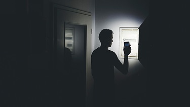 Stromausfall in der Wohnung: Mann leuchtet mit der Taschenlampe in seinem Smartphone in der dunklen Wohnung und überprüft den Sicherungskasten | Bild: picture alliance / CHROMORANGE | Michael Bihlmayer