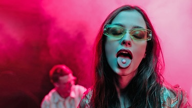 Frau in einer Club-Szene streckt die Zunge heraus, auf der sich eine Pille befindet. | Bild: stock.adobe.com / Lightfield Studios 