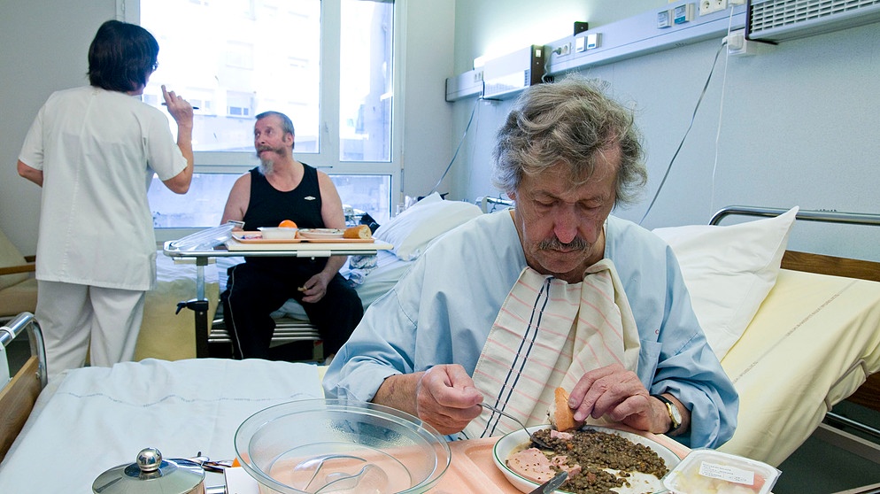 Mann sitzt am Rande eines Krankenhausbettes, vor ihm ein Teller Essen.  | Bild: picture-alliance / BSIP/BELMONTE | BELMONTE