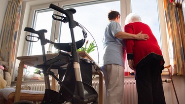 Seniorin mit einer Pflegekraft steht in ihrem Zuhause an einem Fenster | Bild: picture-alliance/dpa