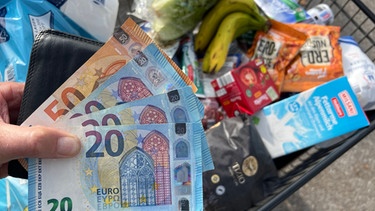 Hand hält aufgefächerte Euro-Geldscheine über einen gefüllten Einkaufswagen | Bild: picture alliance / SvenSimon