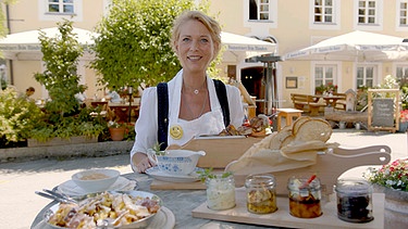 Postwirtin Elisabeth Walch präsentiert Gerichte an einem Holztisch vor ihrem Gasthaus. | Bild: BR, Bewegte Zeiten Filmproduktion GmbH