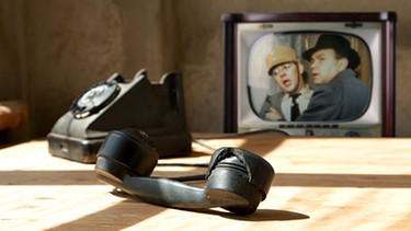 Antikes Telefon auf einem Tisch, im Hintergrund ein Fernseher auf dem ein Szenenbild aus der "Löwengrube" zu sehen ist | Bild: colourbox.com, BR/Tellux-Film GmbH; Montage: BR