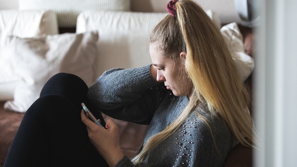 Eine junge Frau sitzt alleine in ihrer Wohnung und hält über ihr Smartphone Kontakt zu ihren Freunden und ihrer Familie. | Bild: BR/Sylvia Bentele