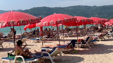 ARCHIV - 11.03.2022, Thailand, Phuket: Touristen liegen unter Sonnenschirmen am Patong Beach. Thailand lockert ab dem 1. Juli die Corona-Reisebestimmungen. Bei der Ankunft müssen nur noch ein Impfausweis oder einen negativen Corona-Test vorlegen werden. Foto: Salinee Prab/AP/dpa +++ dpa-Bildfunk +++ | Bild: dpa-Bildfunk/Salinee Prab