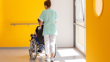Eine Altenpflegerin schiebt eine im Rollstuhl sitzende Person. | Bild: stock.adobe.com/Reddragonfly