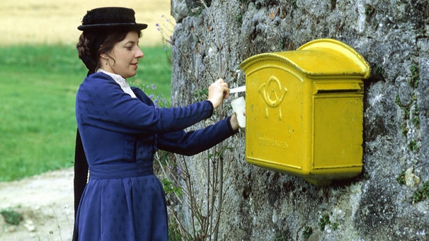 Die Rumplhanni (Monika Baumgartner) wirft einen Brief in einen gelben Briefkasten. | Bild: BR/Foto Sessner