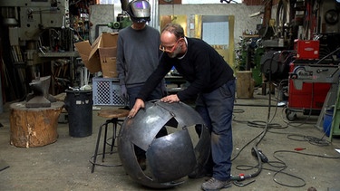 Metallbauer Andreas Pfeiffer in seiner Werkstatt in Hub, der mit Praktikant Shannon Martins, die Segmente aus Stahlblech zu einer Kugel zusammenfügt | Bild: BR