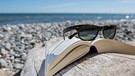 Sonnenbrille liegt auf offenem Buch am Strand.  | Bild: Picture Alliance