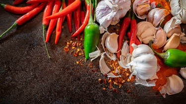 Rote und grüne frische Chilis, ganzer und einzelner Knoblauch, Pfefferkörner und Chilipulver auf einer dunklen Schieferplatte | Bild: 	mauritius images / Robyn Lesley / Alamy / Alamy Stock Photos